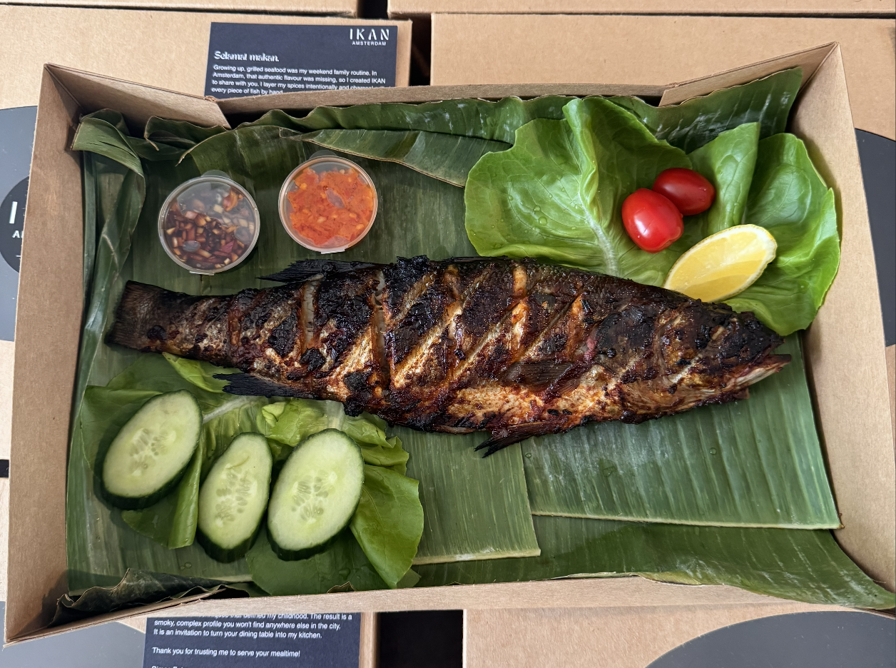 Open box with charcoal-grilled fish, sambal cups, and lalapan on banana leaf